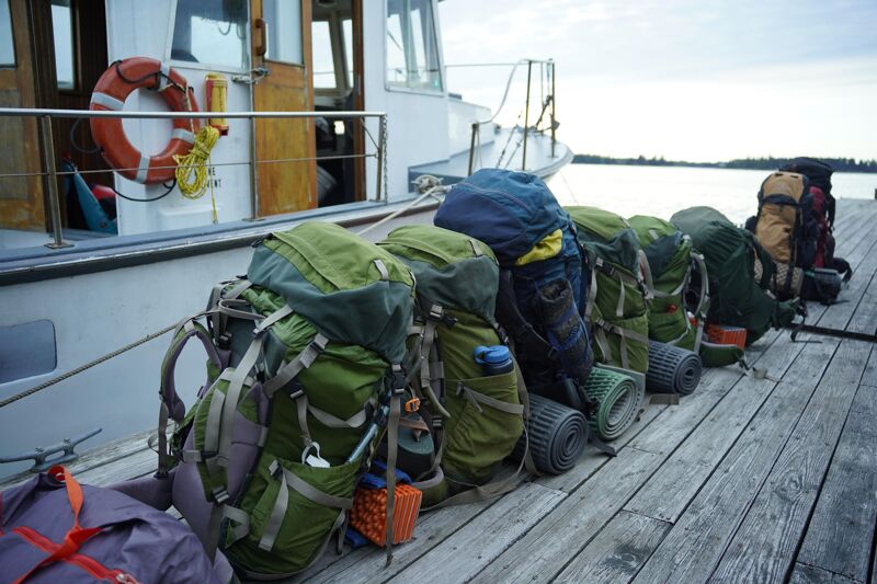 The image shows several backpacks lined up on a wooden dock next to a boat. The backpacks are of various colors and sizes, suggesting a group of people preparing for a trip. The boat is docked, and the scene is set against a backdrop of water and a cloudy sky, indicating an outdoor adventure.